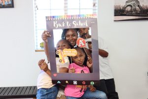 Smiling family poses with a “Back to School” photo frame and playful props during a community event.