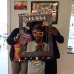 Three smiling participants pose with a colorful “Back to School” photo frame and props during a community event.