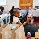 Young girl examines a boxed toy while seated at a table surrounded by gift bags and giveaway items.